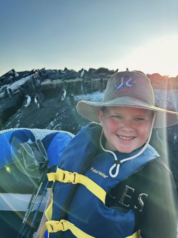 A smiling boy wearing a life vest and a hat smiling at camera with a large rock covered in iguanas behind him.