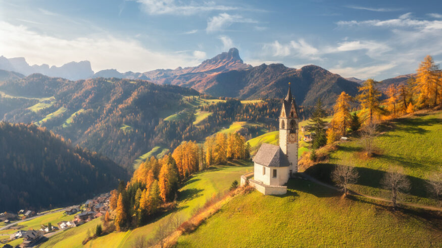 Aerial view of beautiful church, green alpine meadows, orange trees, hills in mountain village at sunset in autumn. Dolomites, Italy. Top view of old chapel, rocks, forest, sky with clouds in fall
