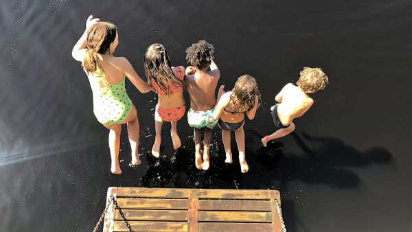 Five small children jumping off a wooden platform into a body of water on a sunny day