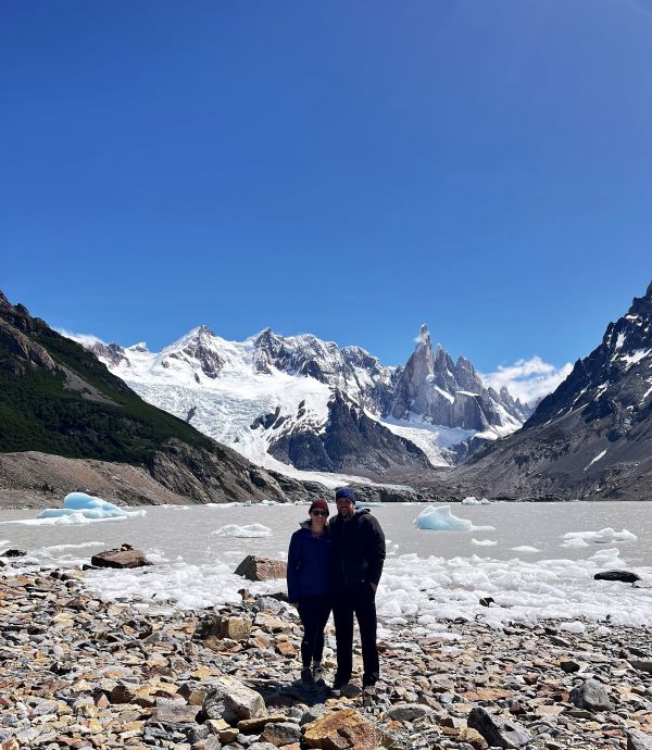 Two hikers standing in front of a lake on a sunny day with the famous Patagonian peak Cerro Torre off in the distance behind.